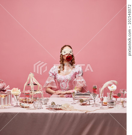 Portrait of charming queen wearing sleep mask and sitting at laying table over pink studio background. Royalty banquet, red rose 101548072