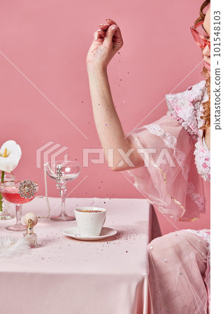 Cropped image of charming blond queen, wearing elegant baroque dress sitting at laid table and preparing coffee with glitter over pink studio background. Cropped image of charming blond queen, wearing elegant baroque dress sitting at laid table and preparing coffee with glitter over pink studio background. 101548103