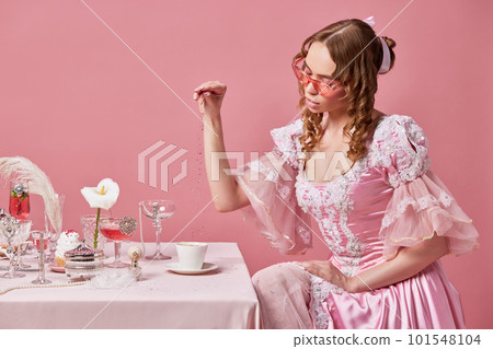 Portrait of charming blond princess, wearing fancy baroque dress sitting at laid table and preparing coffee over pink studio background. Morning royalty breakfast Portrait of charming blond princess, wearing fancy baroque dress sitting at laid table and preparing coffee over pink studio background. Morning royalty breakfast 101548104