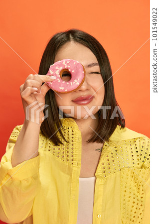 Portrait of young, pretty, asian girl in yellow blouse, holding pink donut, posing against orange studio background 101549022