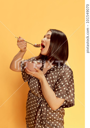 Portrait of beautiful, young, asian girl in stylish silk pajamas, eating cereal with milk against yellow studio background. Delicious breakfast 101549070