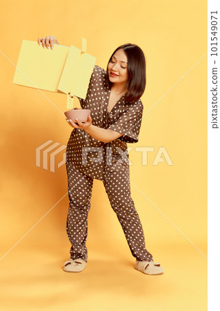Full-length portrait of young, positive, asian girl in comfortable homewear, putting cereal into bowl against yellow studio background. 101549071