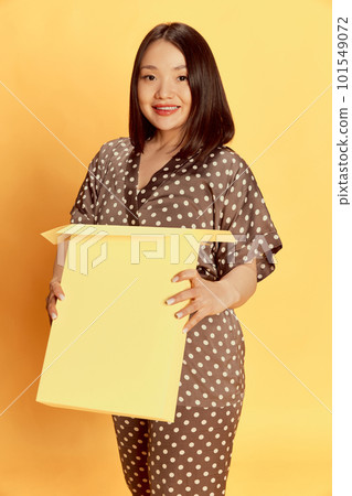 Portrait of pretty, young, asian girl in stylish, comfortable, silk homewear, pyjamas standing against yellow studio background. Smiling 101549072