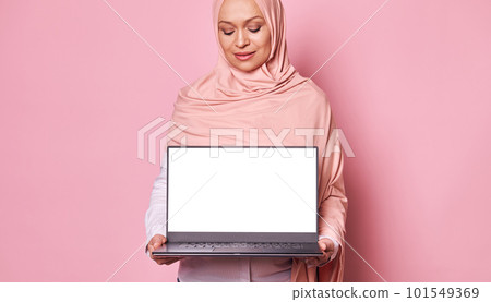 Selective focus on laptop with white blank screen, in the hands of Arab Muslim pretty woman dressed in pink hijab, isolated on pink background. Mockup. Copy advertising space for mobile applications Selective focus on laptop with white blank screen, in the hands of Arab Muslim pretty woman dressed in pink hijab, isolated on pink background. Mockup. Copy advertising space for mobile applications 101549369