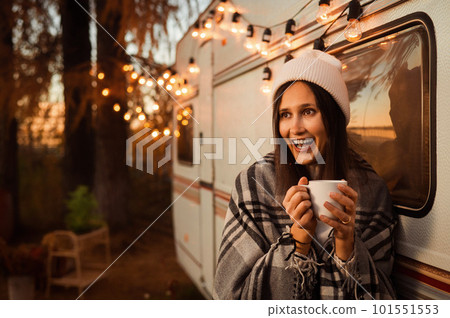 Caucasian woman in a knitted hat wrapped in a plaid and drinks a warming drink outdoors. Travel in a motor home in the fall. Caucasian woman in a knitted hat wrapped in a plaid and drinks a warming drink outdoors. Travel in a motor home in the fall. 101551553