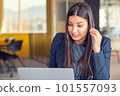 Young Latin Project Manager Putting On Headphones and Working on Laptop Computer in Busy Creative Office Environment. 101557093