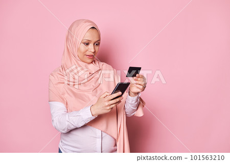 Happy pregnant Arab Muslim woman in hijab, holding credit card and smartphone, shopping online, transferring money via internet banking, ordering baby clothes on mobile apps, isolated pink background Happy pregnant Arab Muslim woman in hijab, holding credit card and smartphone, shopping online, transferring money via internet banking, ordering baby clothes on mobile apps, isolated pink background 101563210