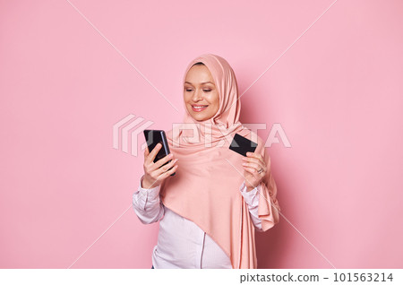 Charming Arab Muslim pregnant woman in pink hijab, holding credit card and smartphone, makes cashless payments via internet mobile banking, isolated background. Online shopping Booking Ordering food 101563214