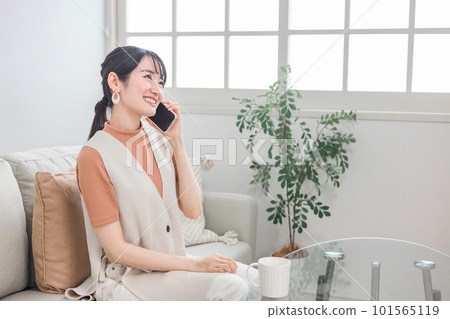Smiling asian woman sitting on sofa and calling friends with smartphone 101565119