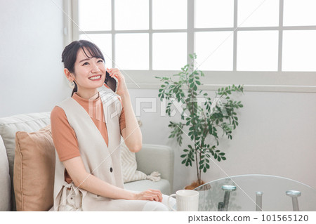 微笑的亞洲女人坐在沙發上,用智能手機給朋友打電話 微笑的亞洲女人坐在沙發上,用智能手機給朋友打電話 101565120