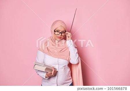Arab Muslim pregnant woman wearing pink hijab, school teacher holds textbooks and pointer, looks at camera through spectacles, expressing misunderstanding and disappointment, isolated pink background 101566878
