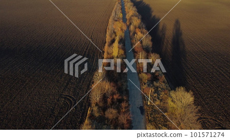Beautiful landscape view old asphalt road with trees and shadows between large plowed agricultural fields of black soil on sunny autumn evening. Flying over dug agrarian fields of earth. Aerial drone Beautiful landscape view old asphalt road with trees and shadows between large plowed agricultural fields of black soil on sunny autumn evening. Flying over dug agrarian fields of earth. Aerial drone 101571274