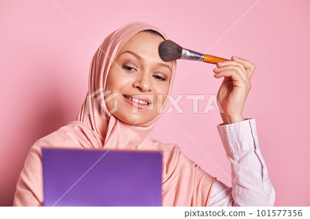 Closeup portrait of a beautiful Middle Eastern Muslim woman in pink hijab, holding palette and using a paintbrush applying makeup foundation and powder on her face, over isolated pink background 101577356