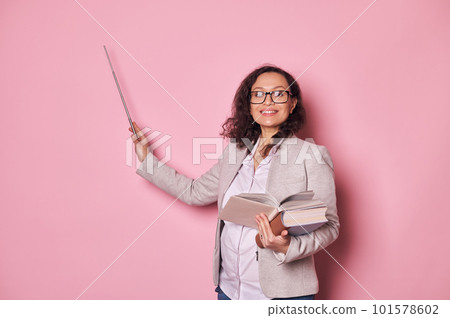 Charming positive pregnant woman, teacher wearing eyeglasses, holding textbooks, smiling, looking at camera, pointing with pointer at copy space on isolated pink background. International Teachers Day 101578602