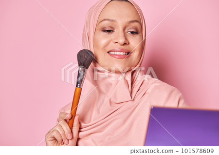 Close Up portrait charming Middle Eastern Muslim woman in pink hijab, smiling looking at her reflection in cosmetic mirror while applying makeup on her face, holding brush, isolated on pink background Close Up portrait charming Middle Eastern Muslim woman in pink hijab, smiling looking at her reflection in cosmetic mirror while applying makeup on her face, holding brush, isolated on pink background 101578609
