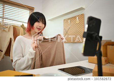 Young woman online seller showing clothes in front of smartphone camera, selling clothes online by streaming Young woman online seller showing clothes in front of smartphone camera, selling clothes online by streaming 101582672