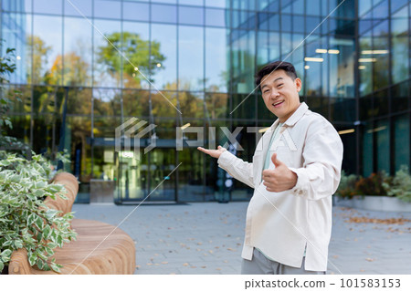 A young Asian man, a businessman, a freelancer, is standing outside an office, a skyscraper, and points to the entrance with his hands. invites with a gesture to come inside, see his project, work. A young Asian man, a businessman, a freelancer, is standing outside an office, a skyscraper, and points to the entrance with his hands. invites with a gesture to come inside, see his project, work. 101583153