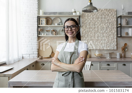 Portrait of a young blogger woman standing at home in the kitchen in an apron and looking at the camera. Blogs about home economics and cooking. Portrait of a young blogger woman standing at home in the kitchen in an apron and looking at the camera. Blogs about home economics and cooking. 101583174