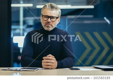 Portrait of an older gray-haired man in glasses of a teacher, trainer, coach who sits seriously in front of the camera and communicates, teaches on a video call. 101583195