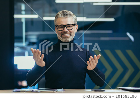 Online psychologist. Senior gray-haired male psychologist, psychotherapist consults with patients remotely via video call. He looks at the camera with a smile, talks amiably. 101583196