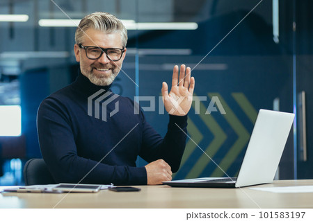 Online psychologist. An older male psychologist, psychotherapist consults with patients remotely via video call. Sitting at the table in front of the laptop, looking and waving at the camera. 101583197