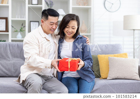 Happy young Asian couple, man and woman sitting on sofa at home. A man gives a woman a red box as a surprise gift. They smile at each other. Happy young Asian couple, man and woman sitting on sofa at home. A man gives a woman a red box as a surprise gift. They smile at each other. 101583214