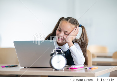 Caucasian girl sits at a desk at school and studies at a laptop. Alarm clock on the table. Caucasian girl sits at a desk at school and studies at a laptop. Alarm clock on the table. 101587416