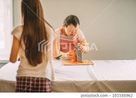 Lifestyle, education. An elderly woman with Down syndrome rubs carrots on a grater with an assistant Lifestyle, education. An elderly woman with Down syndrome rubs carrots on a grater with an assistant 101588962