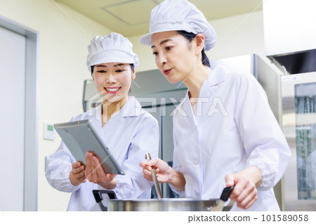 A female cook wearing a white coat prepares food while checking the recipe on a tablet in the kitchen. 101589058