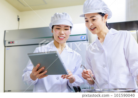A female cook wearing a white coat prepares food while checking the recipe on a tablet in the kitchen. A female cook wearing a white coat prepares food while checking the recipe on a tablet in the kitchen. 101590040
