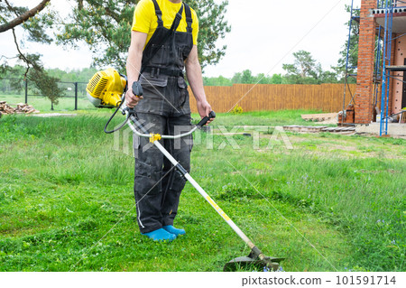 Male gardener mows the green grass of the lawn in the backyard at construction site with a gasoline mower. Trimmer for the care of a garden plot 101591714