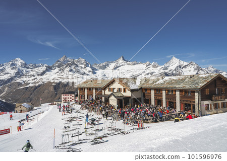 Tourists outside cafe at ski resort chalet with beautiful view of snowcapped mountains and clear blue sky in background at Zermatt, Switzerland, winter holiday travel concept Tourists outside cafe at ski resort chalet with beautiful view of snowcapped mountains and clear blue sky in background at Zermatt, Switzerland, winter holiday travel concept 101596976