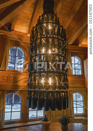 Illuminated chandelier made of empty alcohol bottles hanging on wooden ceiling in alpine chalet with windows in background, luxury travel concept for tourists Illuminated chandelier made of empty alcohol bottles hanging on wooden ceiling in alpine chalet with windows in background, luxury travel concept for tourists 101597003