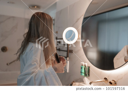 Side view of young blond woman dressed in bathrobe looking in mirror and brushing her hair while standing in modern bathroom at luxurious hotel, luxury vacation concept Side view of young blond woman dressed in bathrobe looking in mirror and brushing her hair while standing in modern bathroom at luxurious hotel, luxury vacation concept 101597316