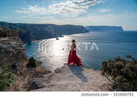 A girl with flowing hair in a long red dress stands on a rock above the sea. The stone can be seen in the sea. Sunny path to the sea from the sun. A girl with flowing hair in a long red dress stands on a rock above the sea. The stone can be seen in the sea. Sunny path to the sea from the sun. 101600781