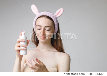 Close up beauty portrait of an attractive young woman apply a cream on her face over white background. Woman has no shirt. Woman has long red hair and wearing bunny ears make-up headband. 101606665