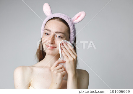 Close up beauty portrait of an attractive young woman wiping her face with a towel isolated over white background. Woman has no shirt. Woman has long red hair and wearing bunny ears make-up headband. 101606669