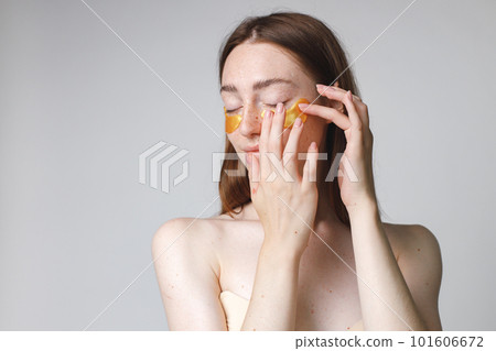 Close up beauty portrait of an attractive young woman applying gel patches on her face isolated over white background. Woman has no shirt. Woman has long red hair. 101606672