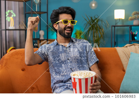 Indian man sits on sofa eating popcorn and watching interesting TV serial, sport game online at home 101606983