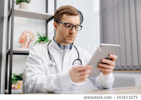 Male Caucasian doctor in white medical uniform filling patient medical history or anamnesis on tablet in office. Doctor consulting client online on tablet in modern clinic, healthcare concept Male Caucasian doctor in white medical uniform filling patient medical history or anamnesis on tablet in office. Doctor consulting client online on tablet in modern clinic, healthcare concept 101611075