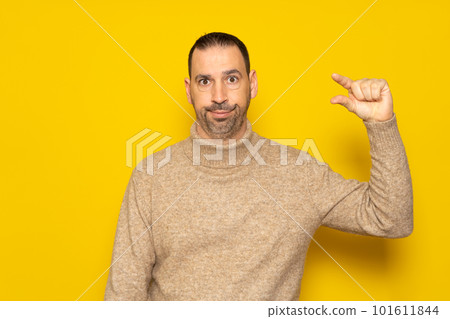 Bearded Hispanic man in his 40s wearing a turtleneck sweater gesturing small or little amount with a disgusted grimace on his face, isolated on yellow studio background 101611844