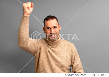 Bearded Hispanic man in his 40s wearing a beige turtleneck raising his fist energetically celebrating a great victory, isolated over gray background 101611845
