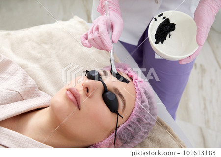 Cosmetologist applying black mask on the face of a beautiful woman for carbon peeling Cosmetologist applying black mask on the face of a beautiful woman for carbon peeling 101613183