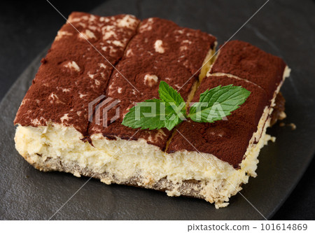 A piece of tiramisu sprinkled with cocoa, on top of a sprig of fresh mint on a black background 101614869