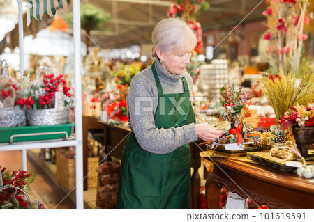 An elderly saleswoman puts Christmas decorations on the shelves in the store 101619391