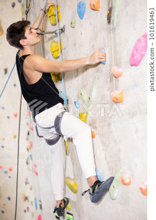 Positive extreme young guy with rope training on concrete climbing rock wall to maintain healthy discipline in indoor adventure park 101619461
