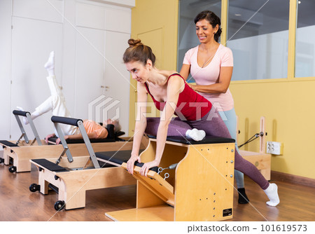 Young woman doing pilates on combo chair with Hispanic female trainer Young woman doing pilates on combo chair with Hispanic female trainer 101619573