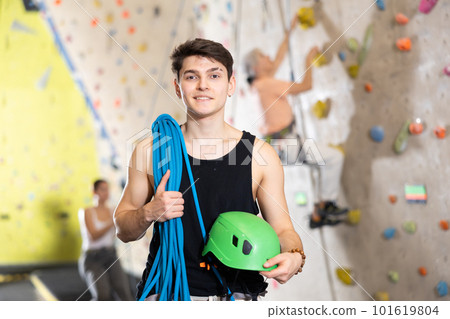 Smiling active young man standing with rope on shoulders and helmet in hand against artificial training climbing wall in adventure park 101619804