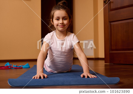 Confident portrait of a beautiful 5-6 years old child, lovely little girl in sportswear, smiling looking at camera, stretching in upward facing dog pose while practicing yoga on a blue fitness mat 101623890
