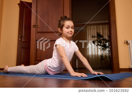 Active little girl wearing comfortable sportswear, practicing yoga, stretching her body Urdhva mukha shvanasana exercise, upward facing dog pose, smiling at camera, working out in homely atmosphere 101623892